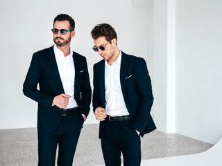 Portrait of two handsome confident stylish hipster lambersexual models. Sexy modern men dressed in black elegant suit. Fashion male posing in white studio interior in sunglasses