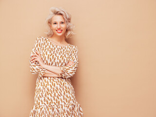 Portrait of young beautiful smiling female in trendy summer dress. Sexy carefree blond woman posing near beige wall in studio. Positive model having fun indoors. Cheerful and happy. Crossed arms