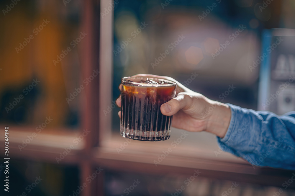 Wall mural hand holding a glass of ice coffee, americano in the afternoon over a cafe glass window background a