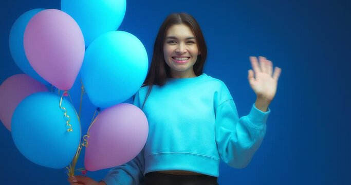 An attractive brunette woman with a beautiful smile, dressed in a blue jumper, holds blue and pink balloons and waves hello on an isolated blue background. Party mood during happy holiday.