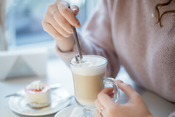 Glass cup with cappuccino and cake on a plate. A woman holding long spoon it in your hand.