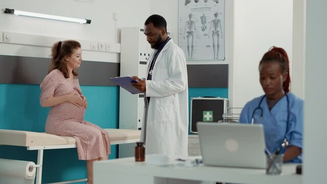 African American Medic Consulting Woman With Pregnancy Belly, Taking Notes On Checkup Files. Male Obstetrician Attending Examination Appointment With Pregnant Patient, Giving Advice.