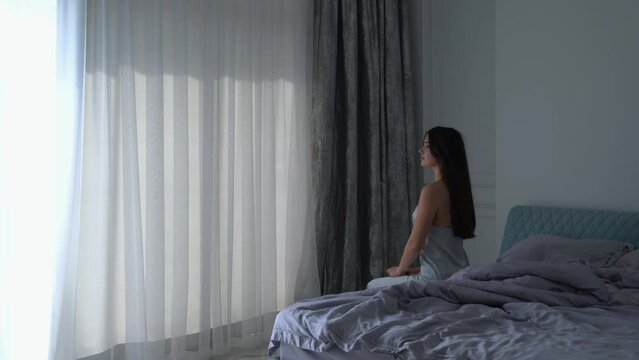 Young woman stand and open white curtains at the window, the morning after waking up in the bedroom of her home.