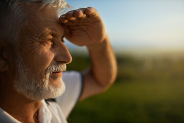 Senior bearded man using hand to protect eyes from sun, smiling outside. Close up view of male tourist squinting from bright light, while looking into distance, during mountain trip. Concept of watch.