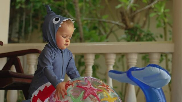 Close-up of a kid plays with rubber inflatable toys outside the room. Infant caucasian toddler in shark costume throwing ball to camera.