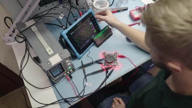 Young electronic developer create prototype of new device and measure electronic circuit parameters. Engineer tests a gadget using a tablet and special equipment. Electronics production in the factory