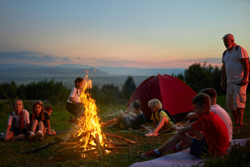 Happy children with adult instructor sitting around campfire in evening. Side view of group of teen tourists resting, having fun, with scenic mountain landscape on background. Concept of travelling.