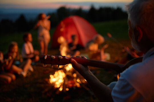Blurred group of teen friends sitting around campfire, having fun in mountains. Crop view of adult bearded man playing guitar and singing to kids, while camping in evening. Concept of camping.