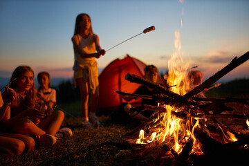 Happy teen girl frying sausage over fire, having fun at campsite at dusk. Side view of group of...