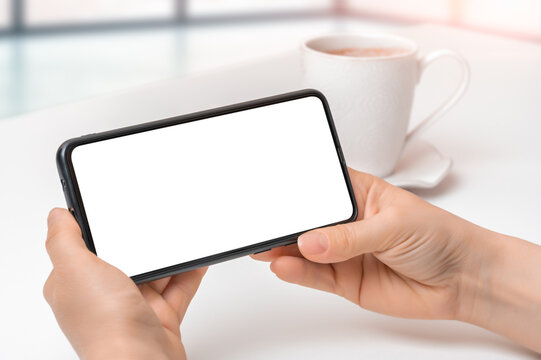 mockup cellphone horizontal position. Hands holding black mobile phone with blank white screen with coffee cup on white table