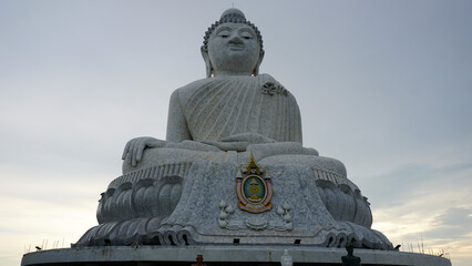 Fototapeta premium The Big Buddha looks straight into the eyes (into the camera). He sits in the lotus position and looks down at people. There is a temple in the center. Gray clouds. Buddha statue in Phuket, Thailand