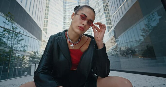 Handsome young woman wearing black jacket skirt stockings and red eyeglasses seductively posing sitting on stairs near glass skyscrapers modern buildings. Gorgeous cheeky urban style fashion model