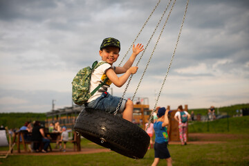 Cheerful kid boy swings on a wheel with chains on a swing in the evening at sunset.