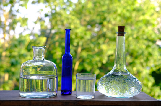 lot of various glass bottles, decanter and vials on the table on wooden table in garden, beautiful blurred natural background with green foliage, concept natural drinks, natural product
