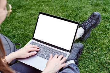 A mockup of a laptop in a girl's lap on the street. A woman works with a laptop in the fresh air in the park, sitting on the lawn. The concept of a remote office. White laptop screen.