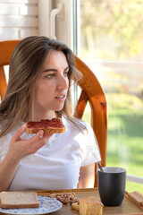 Vertical photo of relaxed young woman sitting having toast and coffee for breakfast as she looks out the window