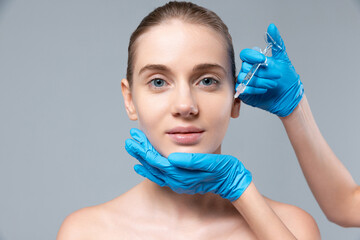 Portrait of young girl doing cosmetology injections on cheeks isolated over grey studio background