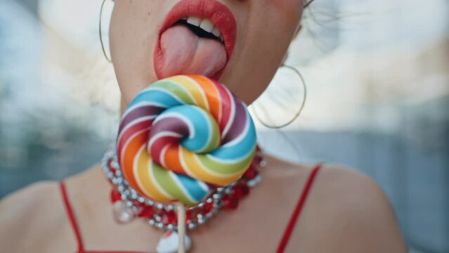 Portrait of gorgeous brunette woman seductively licks big colorful lollipop, stylish teen model wearing cool red eyeglasses posing with multicolored candy, high-rise glass skyscrapers on background