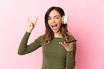 Young caucasian woman isolated on pink background listening music with a mobile making rock gesture