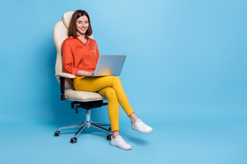 Portrait of attractive cheery girl sitting in armchair using laptop typing project isolated over bright blue color background
