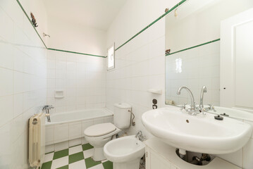 Fototapeta na wymiar Bathroom with white wooden furniture with porcelain sink, frameless mirror on the wall, long tub in the background, green upper border and white and green ceramic floors