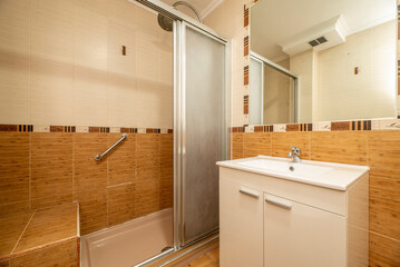 Bathroom with square mirror embedded in the wall, porcelain sink with high-gloss white wooden cabinet, shower tray and imitation wood tiles