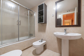 Bathroom with square mirror with wooden frame, porcelain sink of the same material, bathtub with glass screen and ceramic tiles