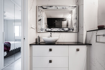 Bathroom with wooden furniture with white drawers, black marble countertop and shelf with metal and...