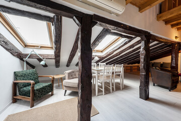 Attic room with wooden dining table and glass top, white chairs, relaxing armchair under a skylight and restored old wooden beams