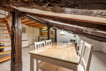 Attic room with wooden dining table and glass top, white chairs and restored vintage wooden beams
