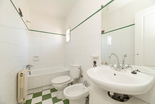 Bathroom with white wooden furniture with porcelain sink, frameless mirror on the wall, long tub in the background, green upper border and white and green ceramic floors