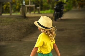 A little curly-haired boy walks in the city in a hat and a yellow T-shirt. A child is playing in an outdoor park. Family vacation, traveler, trip, summer time, side view, life style