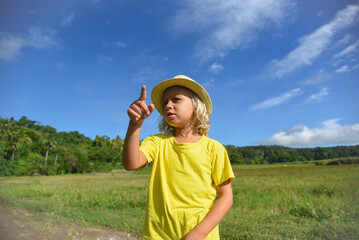 The preschooler shows the direction with his hand. A little curly-haired boy walks in nature in a hat. Vacation, traveler, trip, summer time. Green grass and blue sky in the background, lifestyle