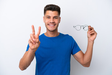 Young caucasian man holding glasses isolated on white background smiling and showing victory sign