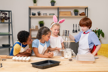 Children pouring dough together, wearing aprons looking at each other with wide smiles while preparing appetizing cookies for their parents