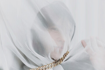 Beautiful woman under veil posing with golden necklace in room. Sensual portrait of young female covered with light fabric. Self care concept. Social isolation and pressure