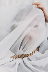 Social isolation and pressure. Woman suffocates under veil, posing with golden necklace. Portrait of young female covered with light fabric. Self care and emotional break concept