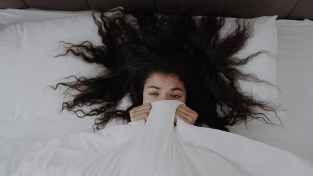 scared funny African American girl lying in bed covering face with blanket, frightened woman covering herself, top view