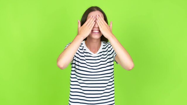 Young brunette woman covering eyes by hands. Surprised to see what is ahead over isolated background. Green screen chroma key