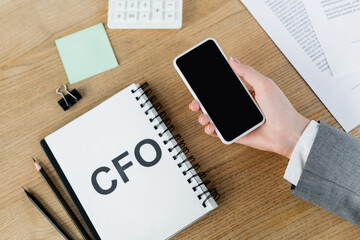 top view of woman holding smartphone with blank screen near notebook with cfo lettering and stationery.