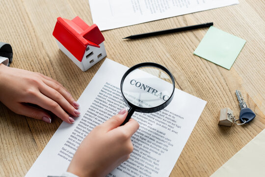 cropped view of tax inspector holding magnifier above contract and house model on desk.