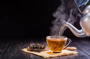Poster Pour a hot herbal teapot into a glass cup, teacup, and dry tea leaves in a wooden spoon and place it on a black wooden table against a dark background. © WIROT