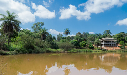 Fototapeta na wymiar Grande lagoa com águas escuras, cercada com muita vegetação e grande construção ao fundo, em área de sítio localizado em Juatuba, Minas Gerais, Brasil.