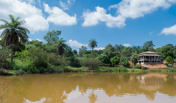 Grande lagoa com águas escuras, cercada com muita vegetação e grande construção ao fundo, em área de sítio localizado em Juatuba, Minas Gerais, Brasil.