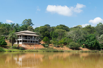 Fototapeta na wymiar Lagoa com águas escuras, cercada com muita vegetação e grande construção ao fundo, em área de sítio localizado em Juatuba, Minas Gerais, Brasil.