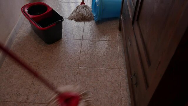 mop is squeezed, wring into the bucket, ready to clean the floor with tiles and colorful curtain on the door in the background. a second mop overturned to the side.