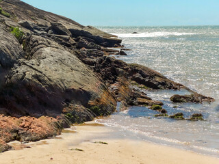 Linda praia, com grandes rochas, banhada pelas ondas, muita vegetação e montanhas em volta e lindo céu azul localizada em Vitória no estado do Espírito Santo, Brasil.