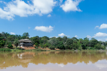 Fototapeta premium Linda lagoa com águas escuras, cercada com muita vegetação e grande construção ao fundo, em área de sítio localizado em Juatuba, Minas Gerais, Brasil.