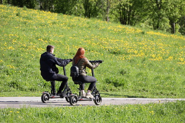 Guy and girl ride electric scooters with seats in summer park on background of green hill with...