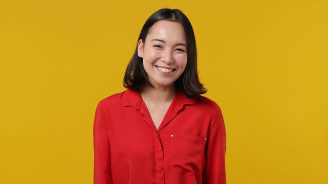Smiling charming vivid overjoyed happy young woman of Asian ethnicity 20s years old wears red shirt stand up appear looking camera wink eye blink isolated on plain yellow background studio portrait
